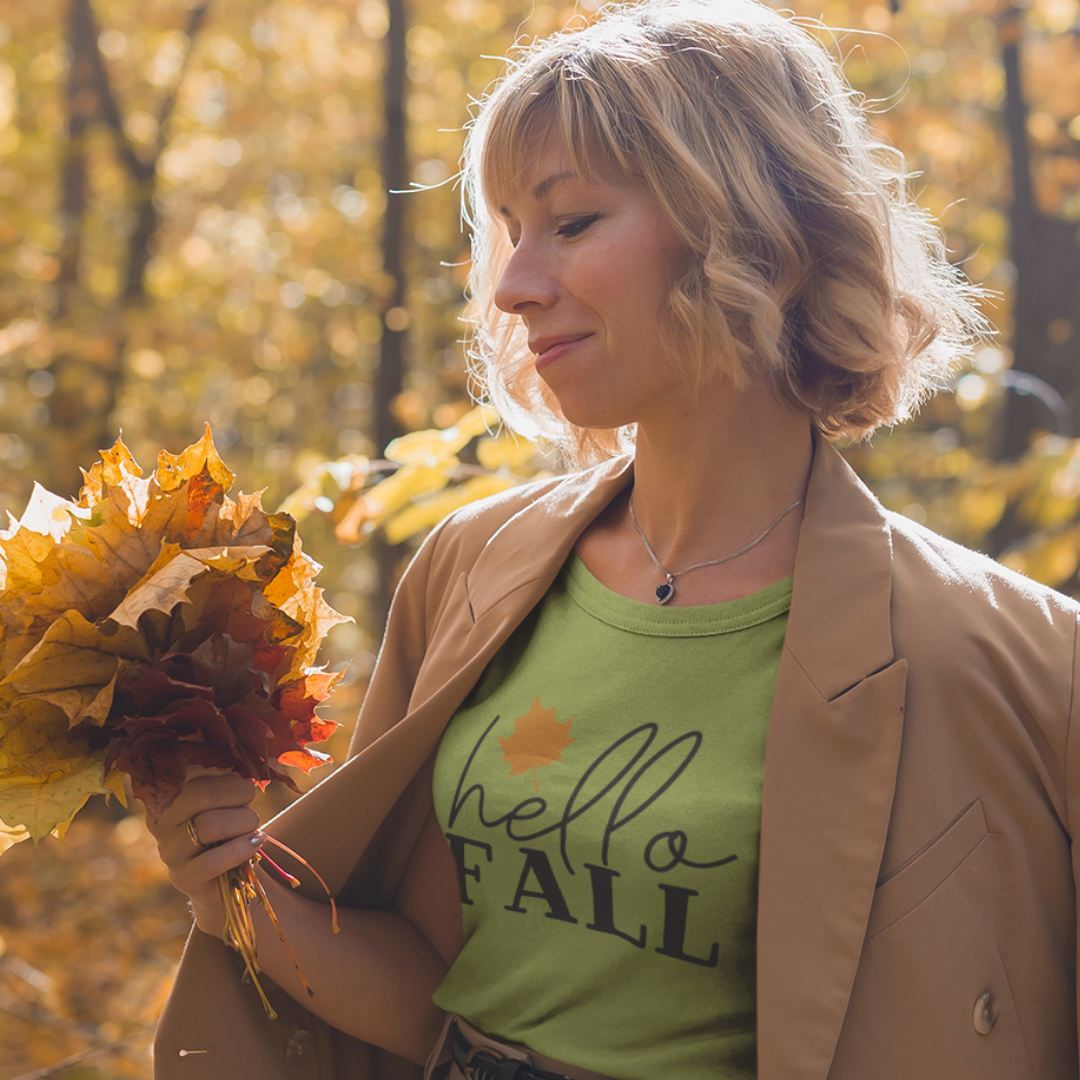 Woman holding autumn leaves wearing a Hello Fall olive/sage green t- shirt in a forest setting