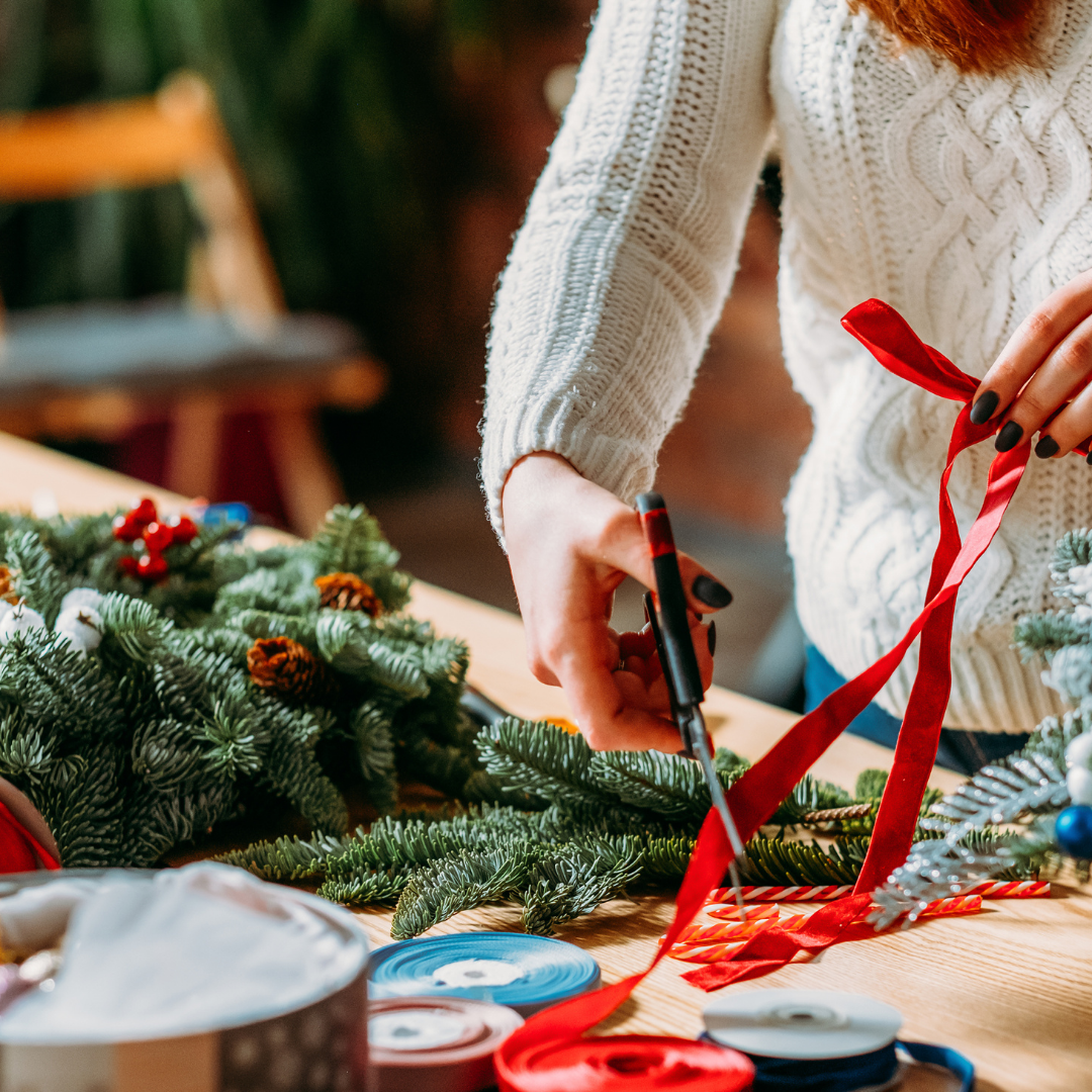 Person decorating a Christmas wreath with red ribbon and scissors.
