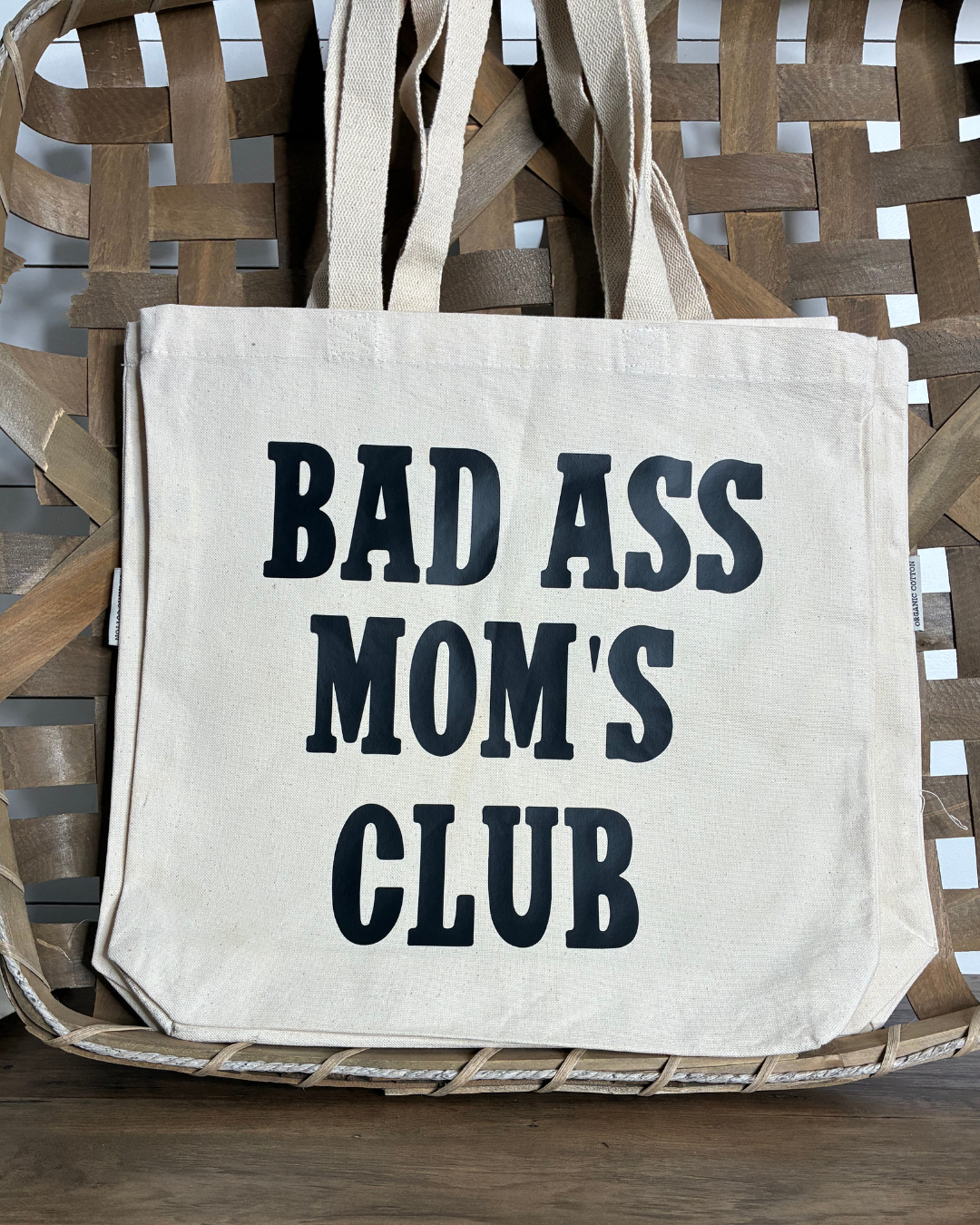 Sustainable cream canvas tote bag with bold black "Bad Ass Mom's Club" text, shown in a rustic woven basket.