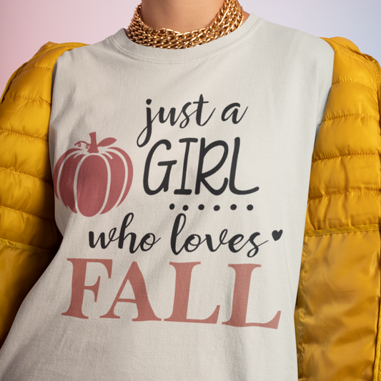 A woman wearing a sand/neutral shirt with Just a girl who loves fall text and a yellow puffer jacket. Available in fall colors and sizes