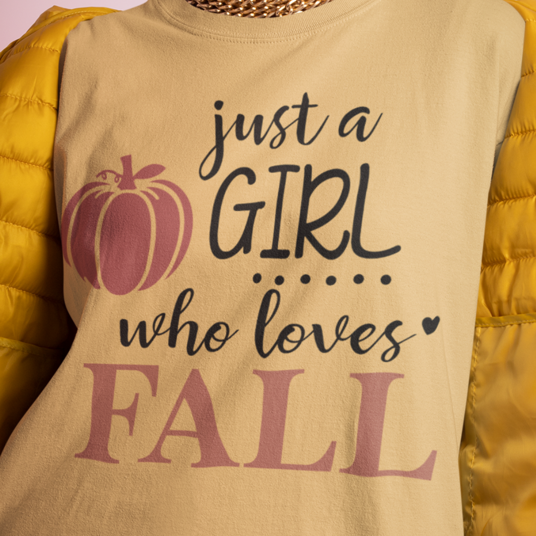 Person wearing a sand/neutral shirt with Just a girl who loves fall text and a yellow puffer jacket. Available in fall colors and sizes