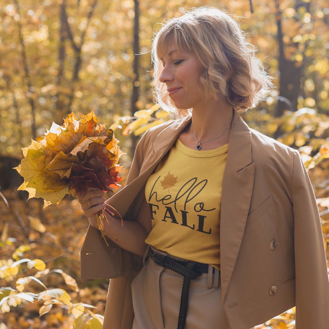 Woman in a mustard Hello Fall T-shirt holding autumn leaves in a forest.