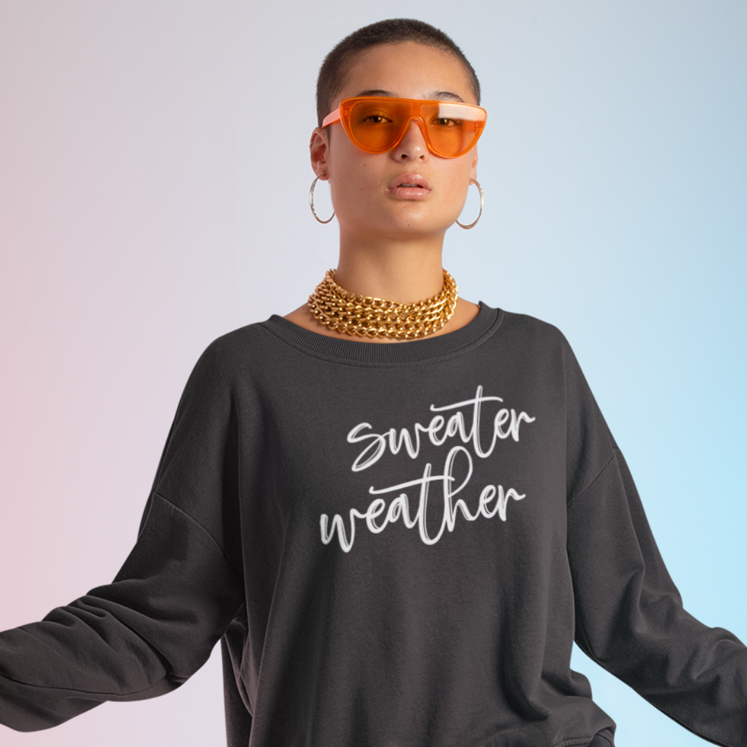 Minimalist Sweater Weather Sweatshirt in a black color with white font, folded on a bed of fall leaves.