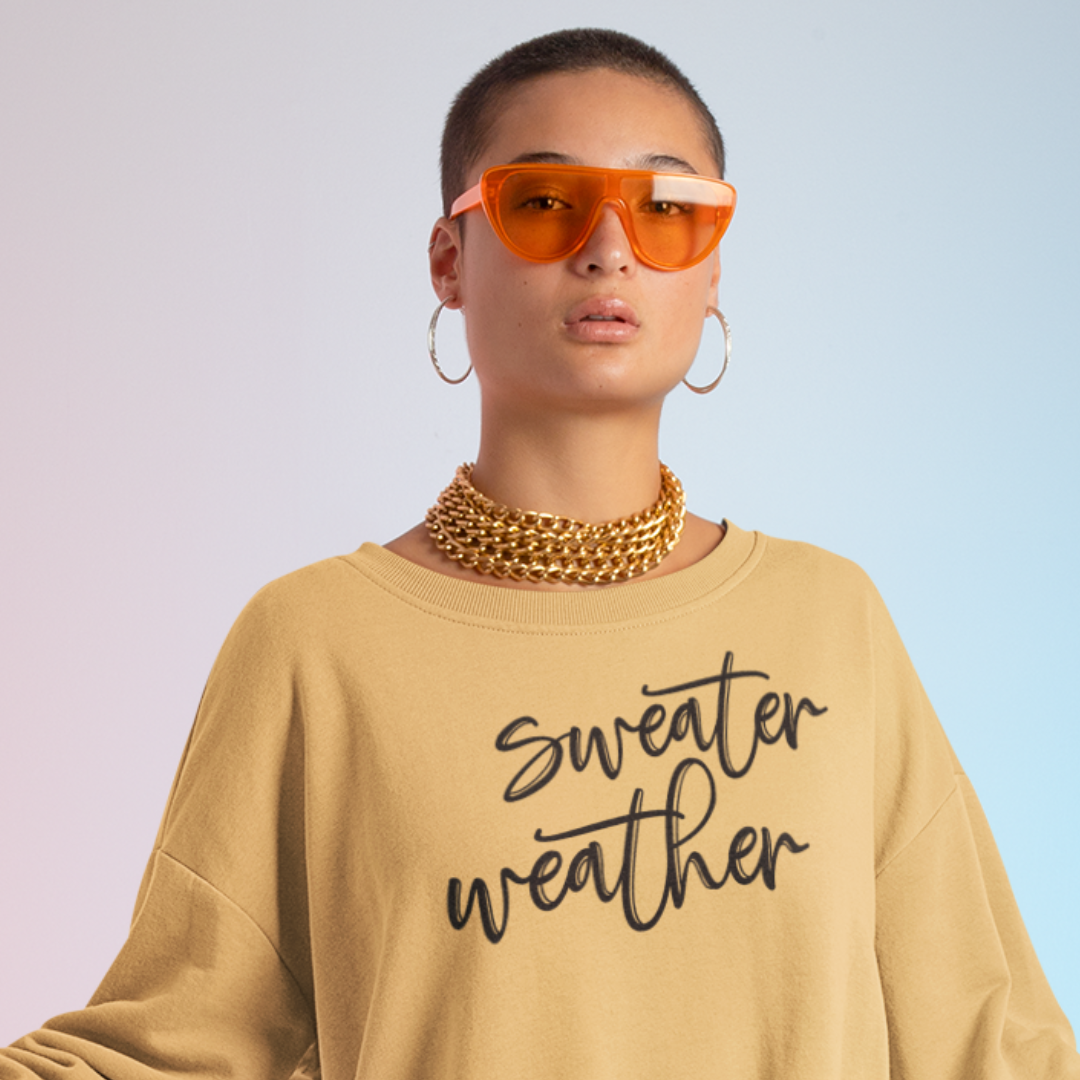 Minimalist Sweater Weather Sweatshirt in mustard tone white font, folded on a bed of fall leaves.