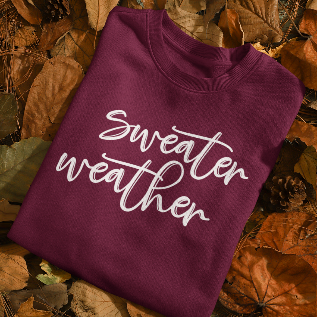 Minimalist Sweater Weather Sweatshirt in a plum/marron tone with white font, folded on a bed of fall leaves.