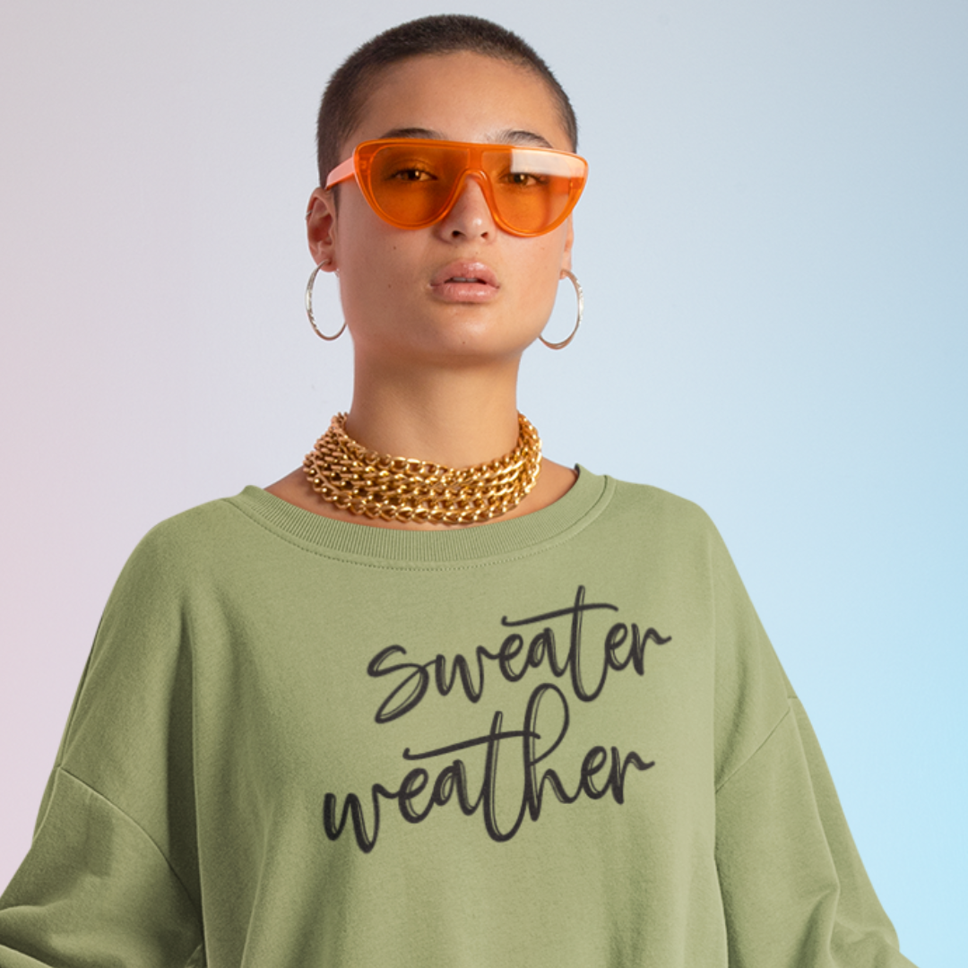 Minimalist Sweater Weather Sweatshirt in olive green tone white font, folded on a bed of fall leaves.
