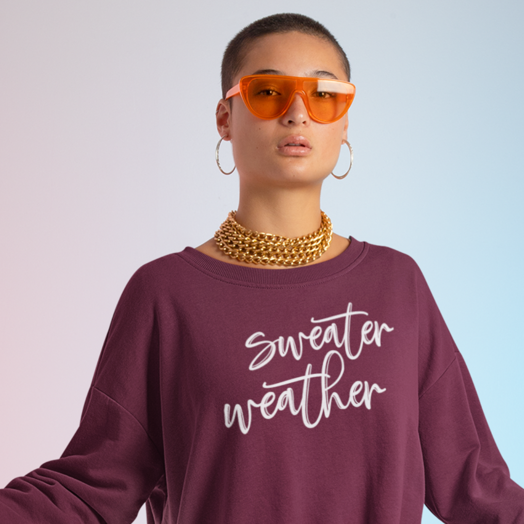 Minimalist Sweater Weather Sweatshirt in plum/maroon tone white font, folded on a bed of fall leaves.