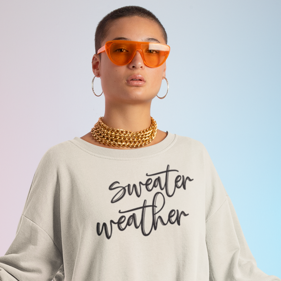 Minimalist Sweater Weather Sweatshirt in sand tone white font, folded on a bed of fall leaves.