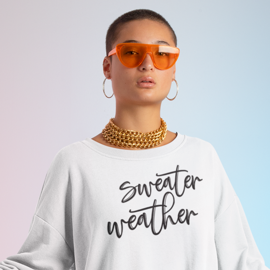 Minimalist Sweater Weather Sweatshirt in white tone white font, folded on a bed of fall leaves.