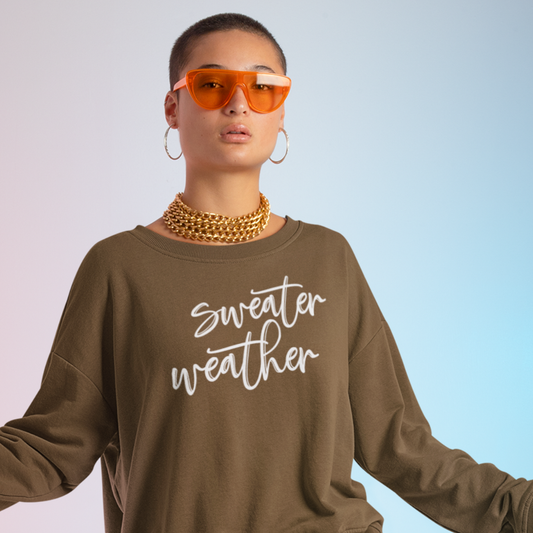 Minimalist Sweater Weather Sweatshirt in a brown tone with white font, folded on a bed of fall leaves.