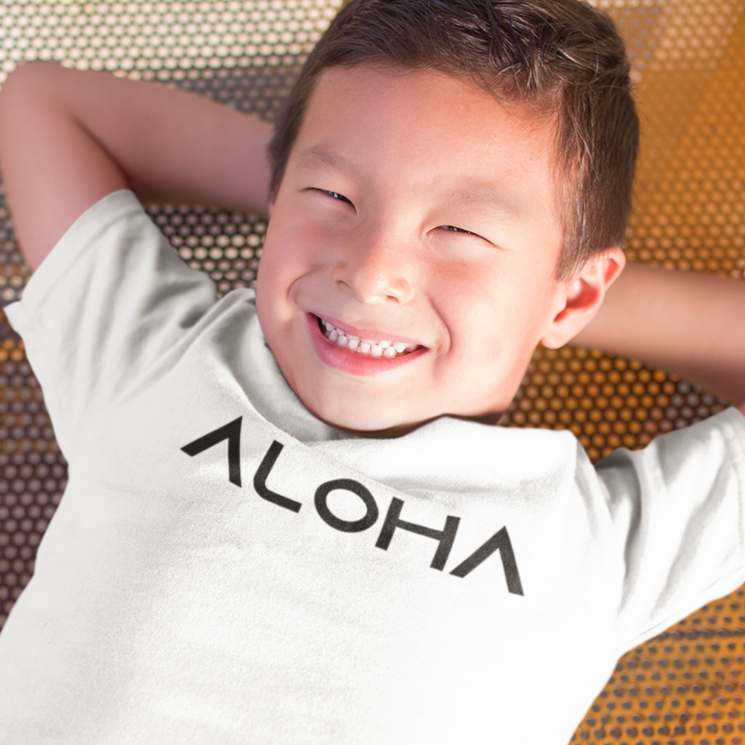 A young boy smiling while wearing a white minimalist Kids Aloha Shirt from Simply Aloha On The Go.
