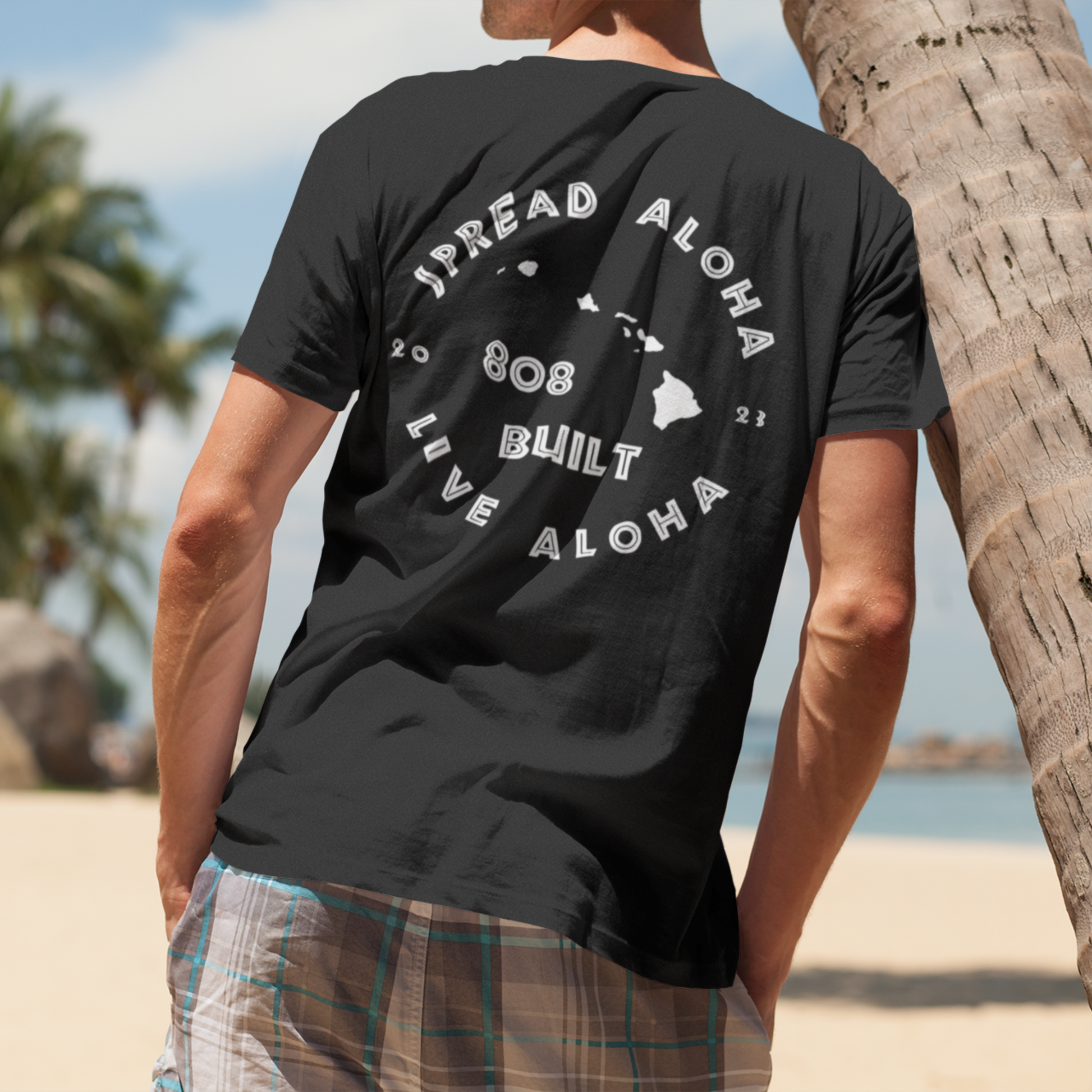 Back view of a man on a beach wearing a black 808 Built Hawaii t-shirt with a white circular graphic that reads Spread Aloha Live Aloha 808 Built

