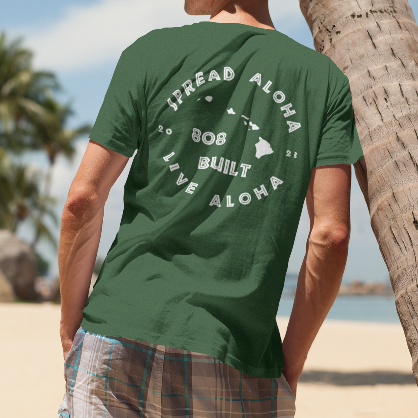 Back view of a man on a beach wearing a green 808 Built Hawaii t-shirt with a white circular graphic that reads Spread Aloha Live Aloha 808 Built

