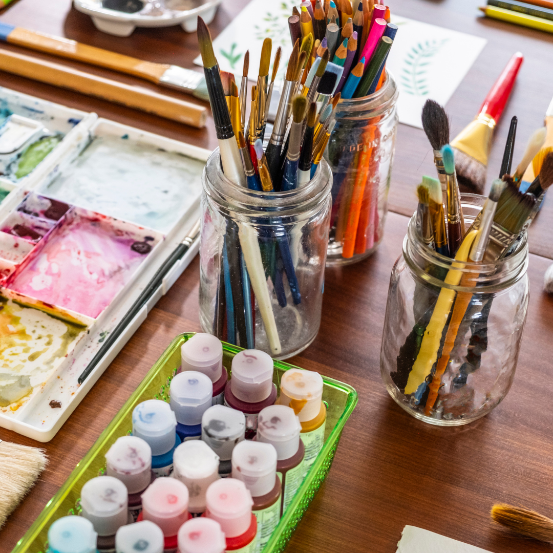 Art supplies including paintbrushes in jars and paint tubes on a wooden table.