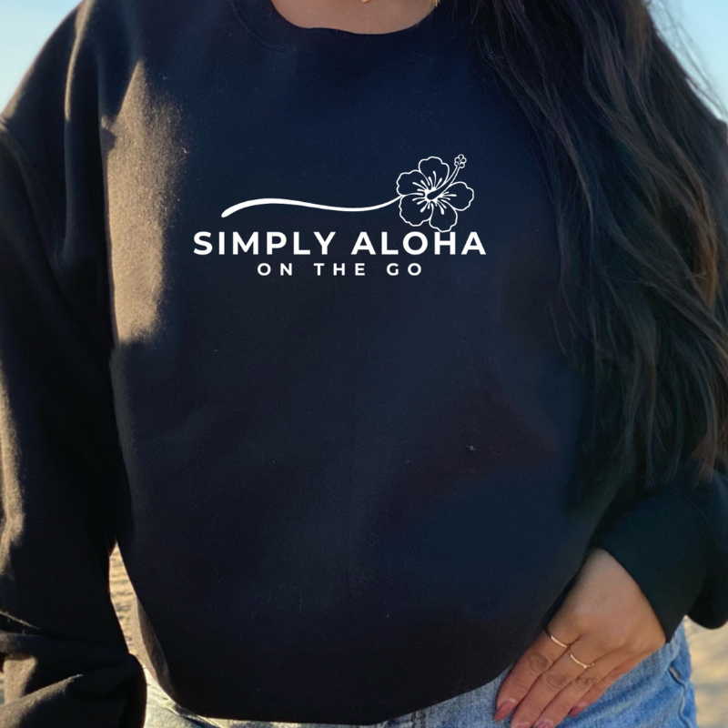 A closeup of a woman wearing her Simply Aloha On The Go Sweatshirt in black color at the beach – Island-Inspired Crewneck in Black with  White font

