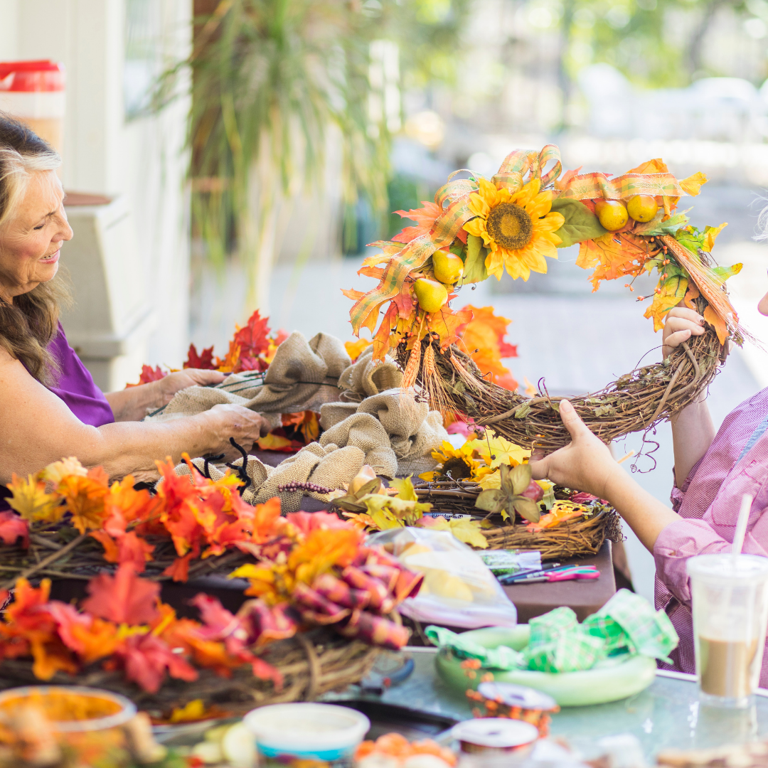 Two women working on a craft project with decorative items including a sunflower wreath.