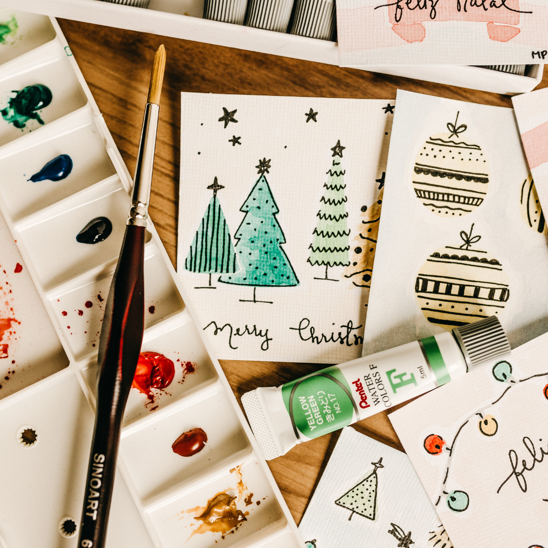 Art supplies including watercolor paints, brushes, and Christmas-themed cards on a wooden surface.