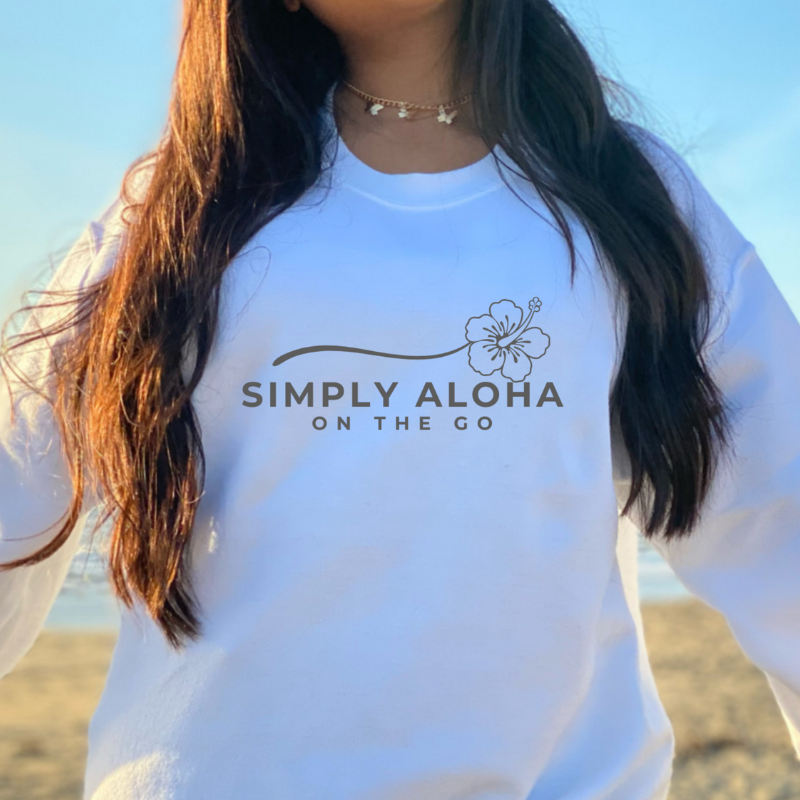 A smiling woman by the beach wearing a white custom crewneck sweatshirt featuring a clean black brand logo that reads Simply Aloha On The Go.