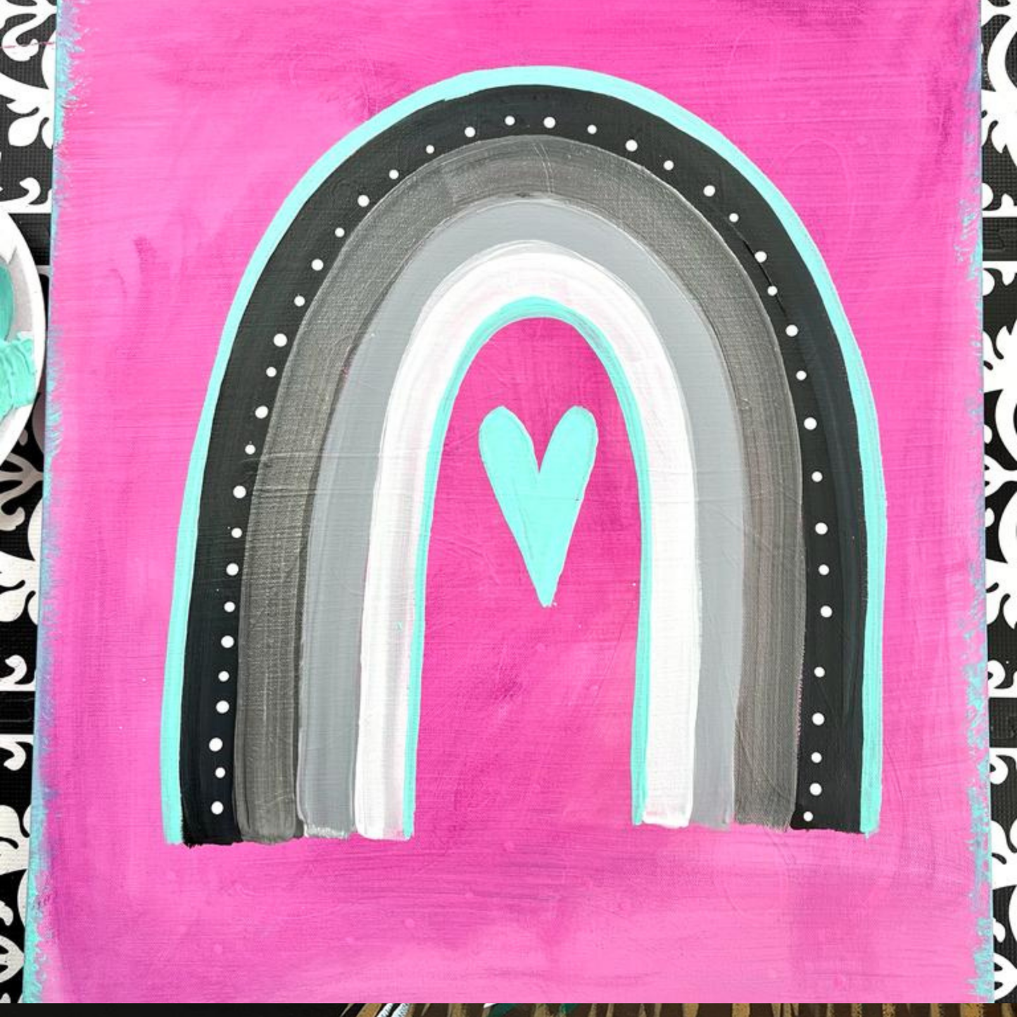 A grey and white boho-style rainbow with a small teal heart in the center on a bright pink background.