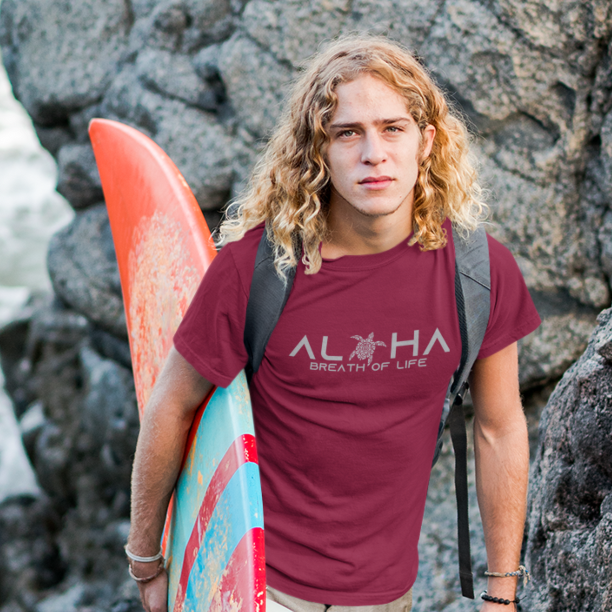 Man wearing a maroon Men's Breath of Life T-Shirt, Hawaiian-inspired tee featuring a honu sea turtle while holding a colorful surfboard.