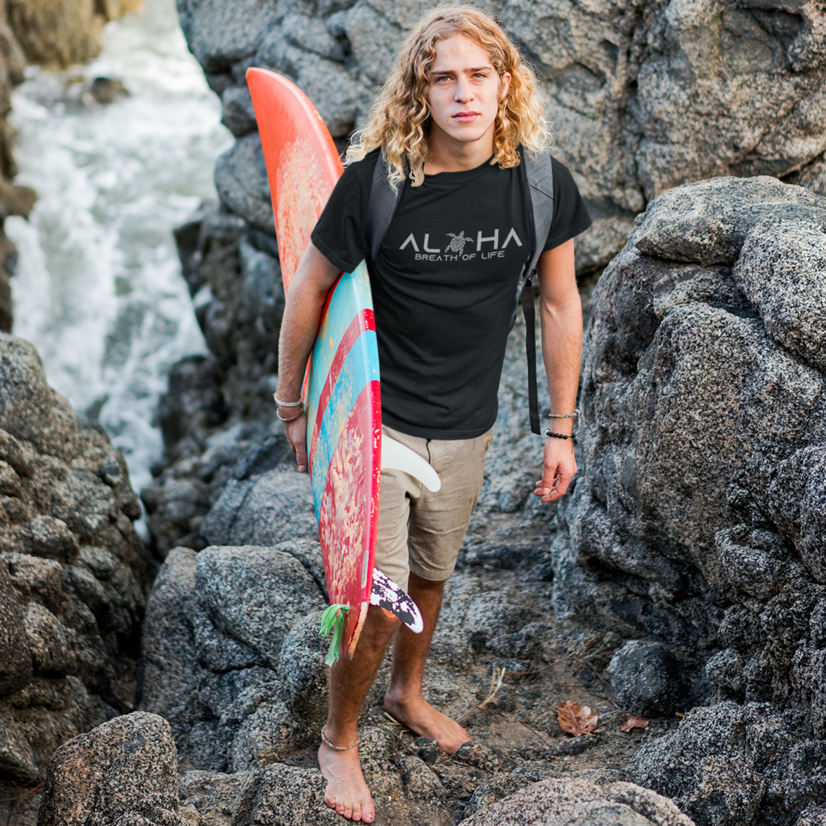 Man wearing ablack Men's Breath of Life T-Shirt, Hawaiian-inspired tee featuring a honu sea turtle while holding a colorful surfboard.