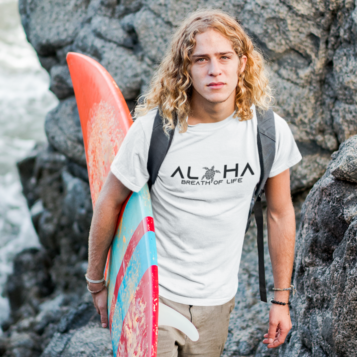 Man wearing a white Men's Breath of Life T-Shirt, Hawaiian-inspired tee featuring a honu sea turtle while holding a colorful surfboard.
