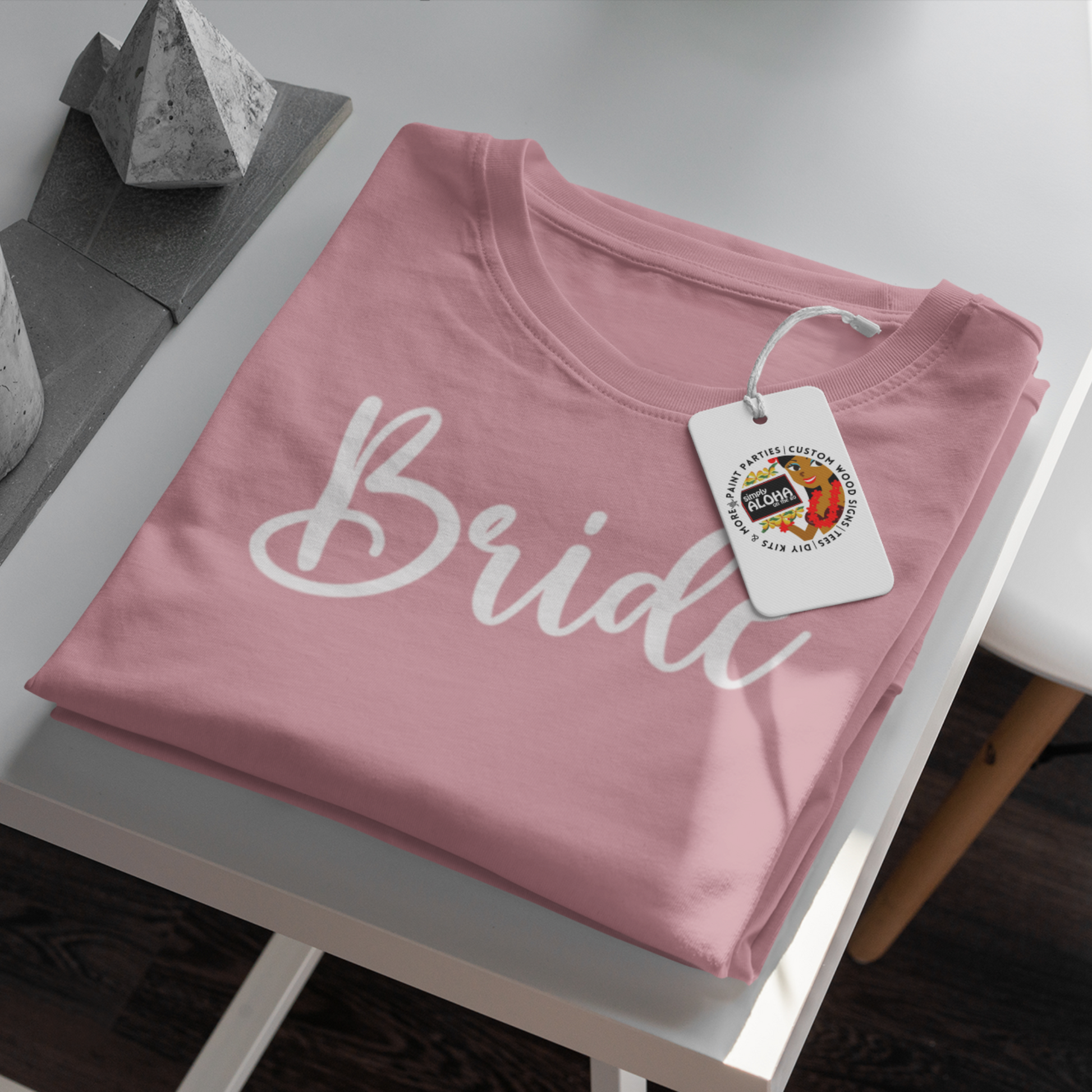 A closeup look of a clean and chic pink t-shirt with the word "Bride" printed in a subtle, elegant white font. The shirt is a must-have for a bridal shower or bachelorette party