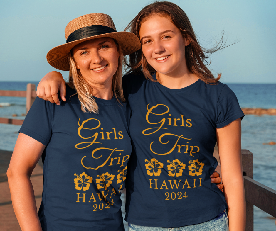 Two women standing by the ocean wearing matching navy blue custom girls trip t-shirts with white lettering.