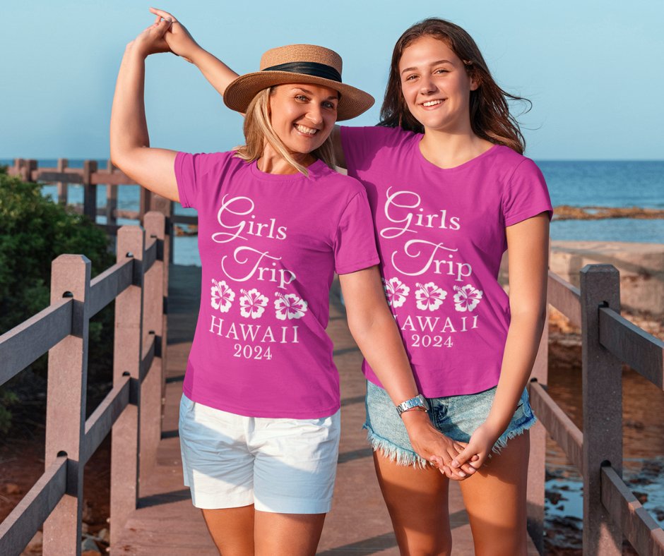 Two women smiling outdoors by the water wearing matching custom girls trip t-shirts in pink that reads Wear The Aloha Live The Vibes.