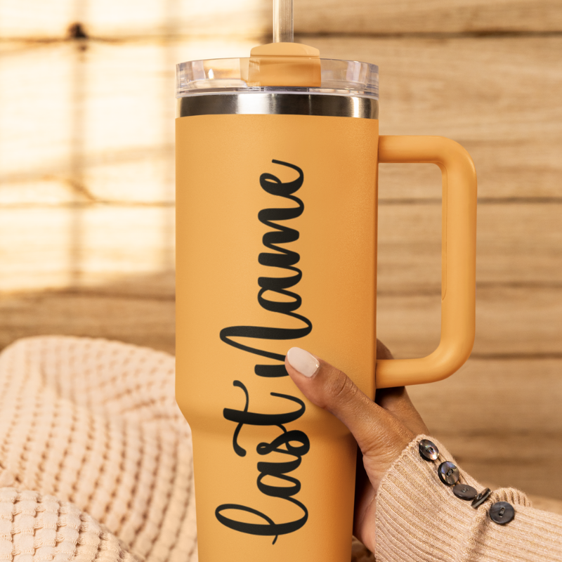 A close-up of a saffron tumbler held by a hand against a wooden background with a custom black vinyl decal featuring the last name you'd like to add. The decal is applied smoothly to the side of the cup by Simply Aloha On The Go