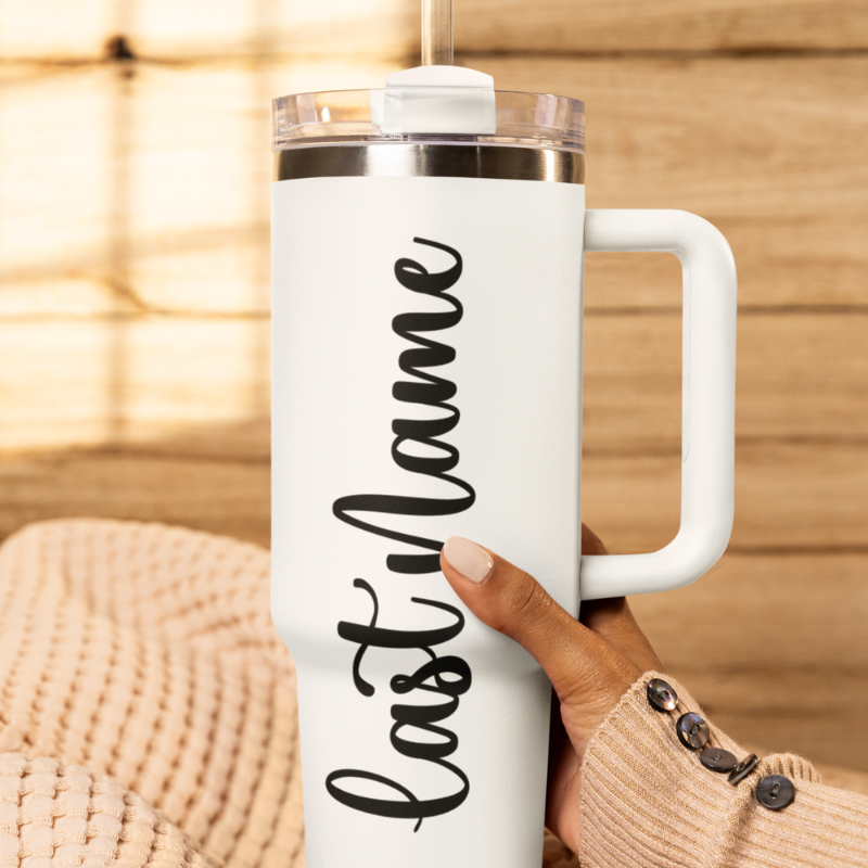 A close-up of a white tumbler held by a hand against a wooden background with a custom black vinyl decal featuring the last name you'd like to add. The decal is applied smoothly to the side of the cup by Simply Aloha On The Go