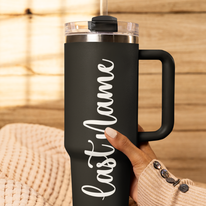 A close-up of a black tumbler held by a hand against a wooden background with a custom white vinyl decal featuring the last name you'd like to add. The decal is applied smoothly to the side of the cup by Simply Aloha On The Go