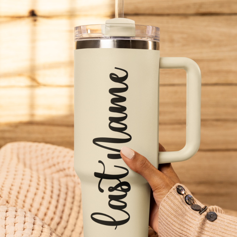 A close-up of a sand tumbler held by a hand against a wooden background with a custom black vinyl decal featuring the last name you'd like to add. The decal is applied smoothly to the side of the cup by Simply Aloha On The Go
