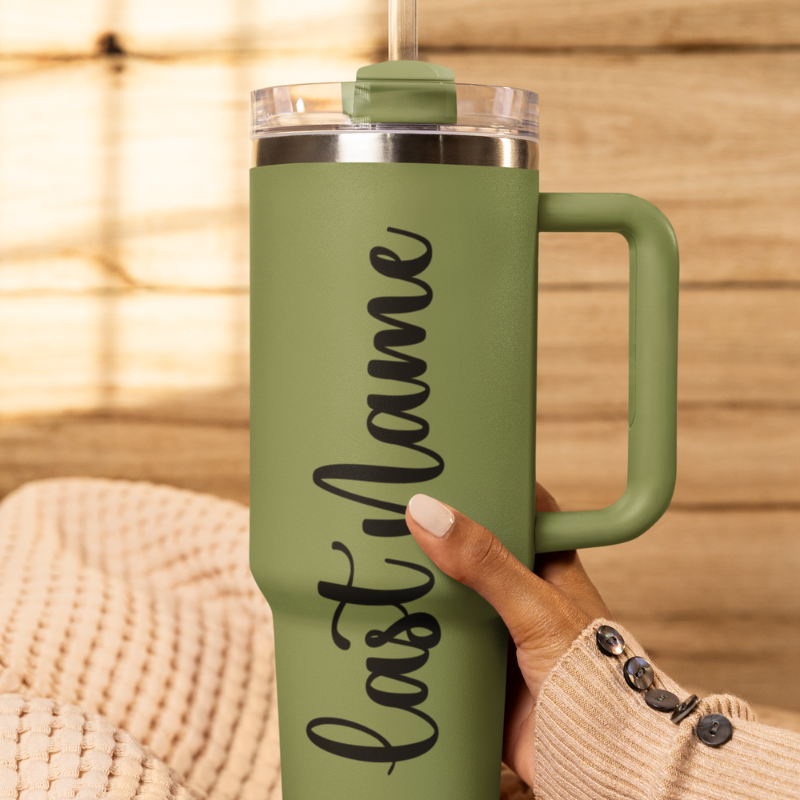 A close-up of a sage green tumbler held by a hand againts a wooden background with a custom black vinyl decal featuring the last name you'd like to add. The decal is applied smoothly to the side of the cup.
