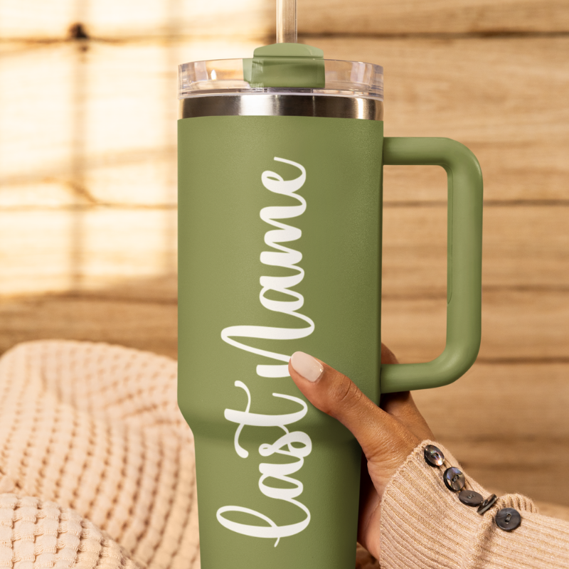 A close-up of a sage green tumbler held by a hand againts a wooden background with a custom white vinyl decal featuring the last name you'd like to add. The decal is applied smoothly to the side of the cup.