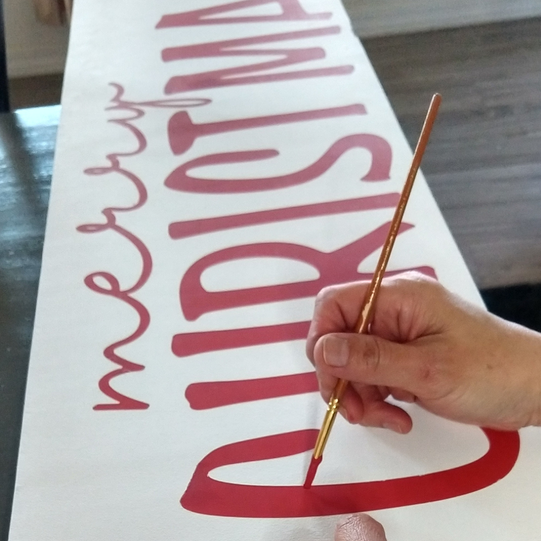 Hand-stenciling a personalized  Merry Christmas phrase porch sign, holiday front porch decor. Custom farmhouse Christmas porch sign with a personalized "Merry Christmas" greeting in red font on a white background on weathered wood.