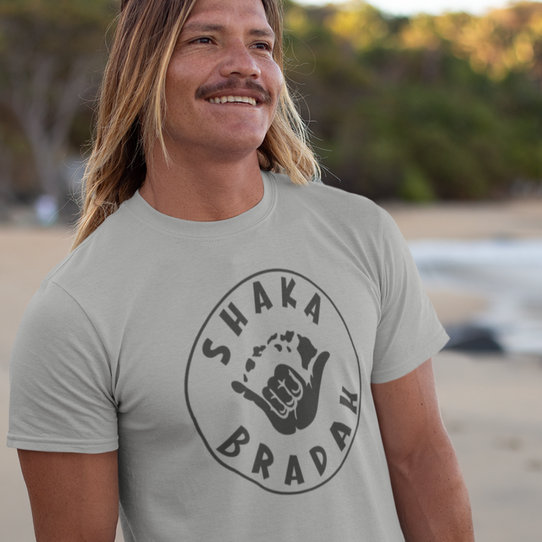 A man wearing a grey "Shaka Bradah"t-shirt, black font with a graphic of a hand making the shaka sign.