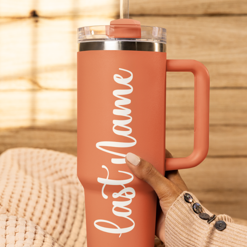 A close-up of a salmon tumbler held by a hand against a wooden background with a custom white vinyl decal featuring the last name you'd like to add. The decal is applied smoothly to the side of the cup by Simply Aloha On The Go