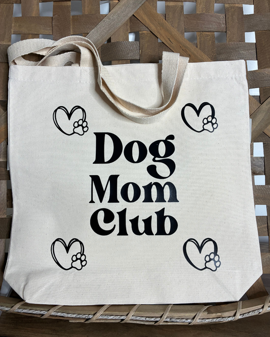 Dog Mom Club Tote Bag in natural cotton canvas featuring Dog Mom Club in lettering and heart illustrations, displayed on a wooden chair.