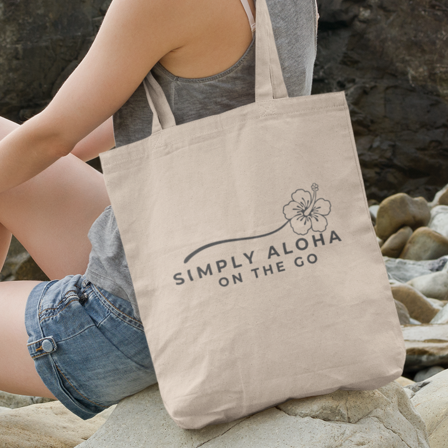 Person sitting on rocks with a beige tote bag featuring Simply Aloha On The Go text with hibiscus design.