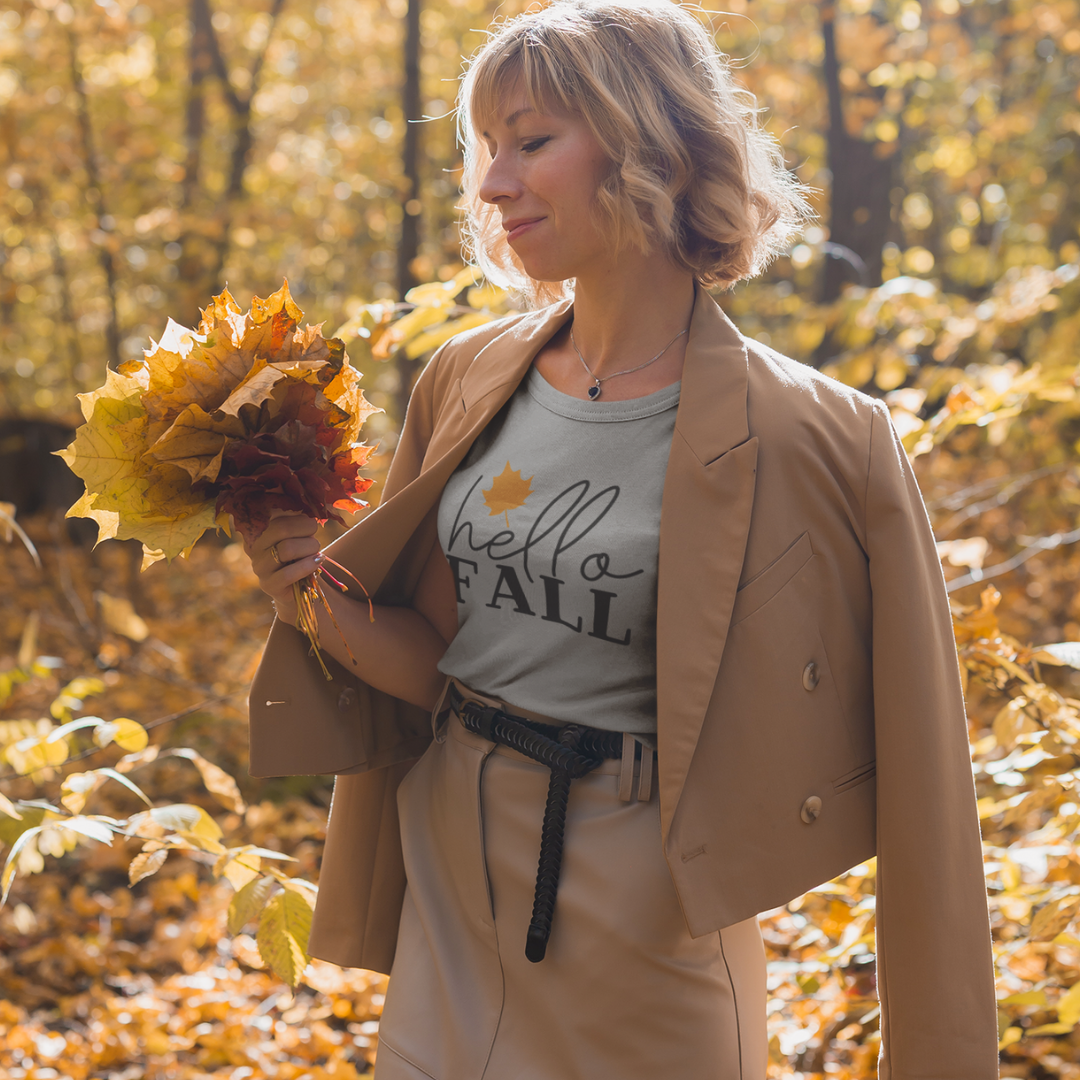 Woman in a beige coat holding autumn leaves with hello FALL shirt in a forest.