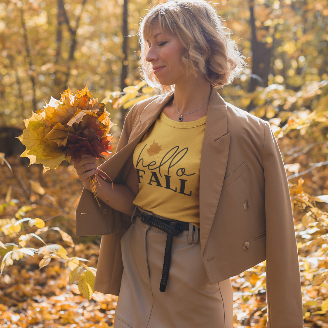 Woman in a yellow hello FALL shirt holding autumn leaves in a forest.