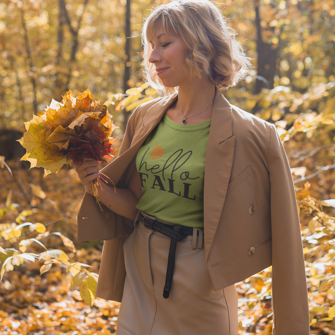 Woman in a green hello FALL shirt holding autumn leaves in a forest.