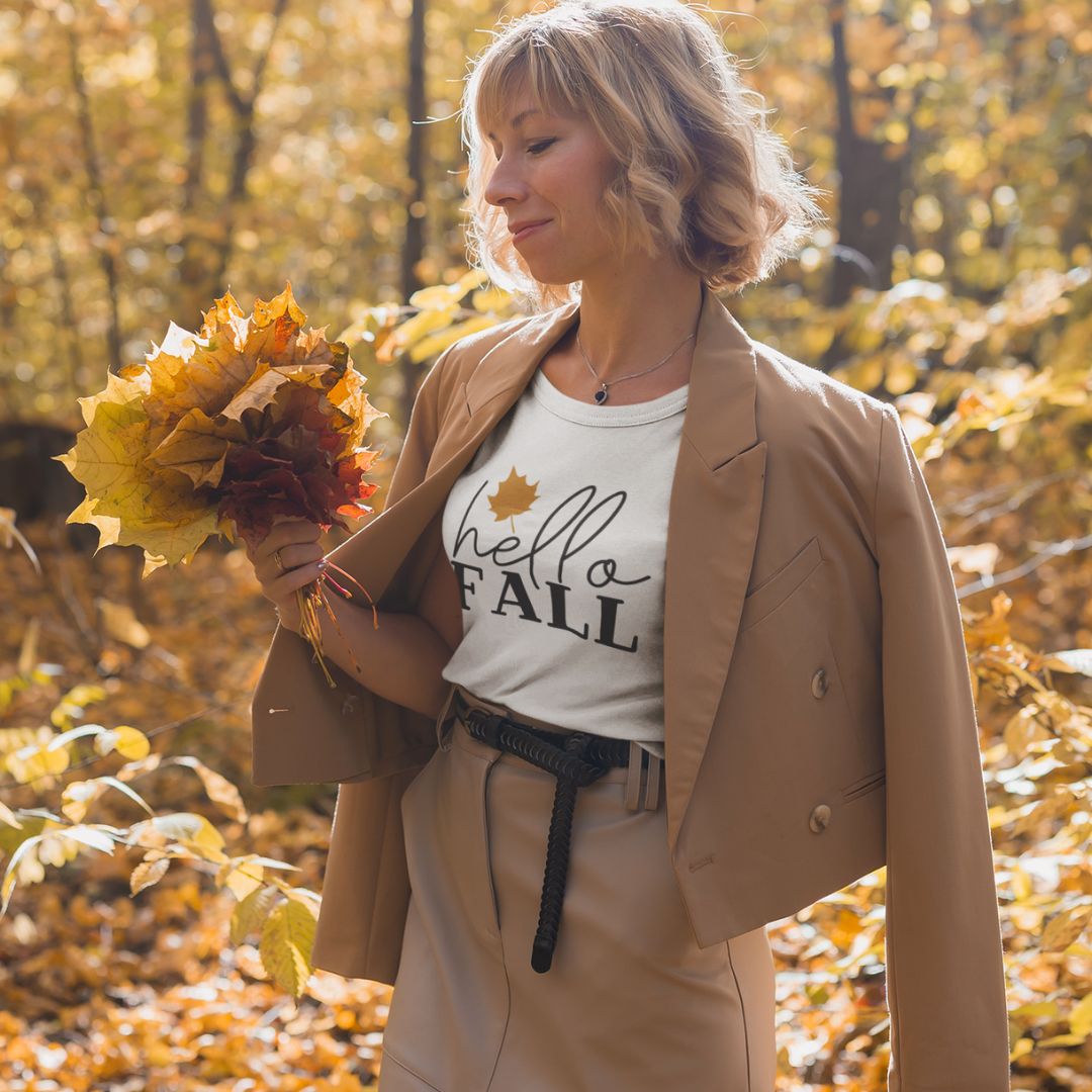 Woman in a beige coat holding autumn leaves with hello FALL shirt in a forest.