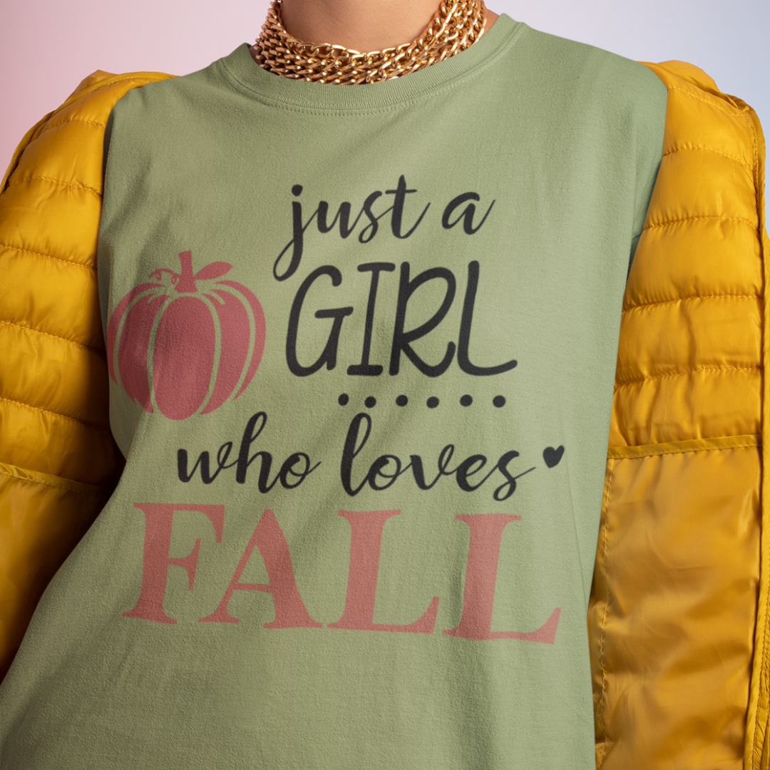 A woman wearing a olive green Just A Girl Who Loves Fall graphic t-shirt featuring a minimalist text design and a small pink pumpkin illustration.