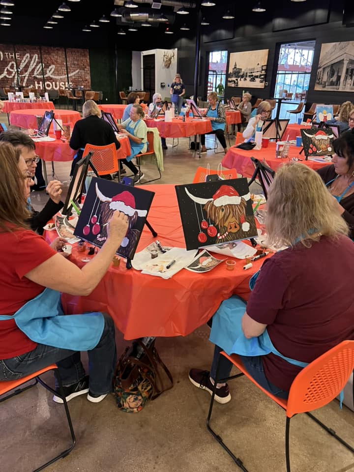 People sitting at tables with art supplies and paintings in a room labeled The Coffee House painting the holiday famrhouse Highland cow.