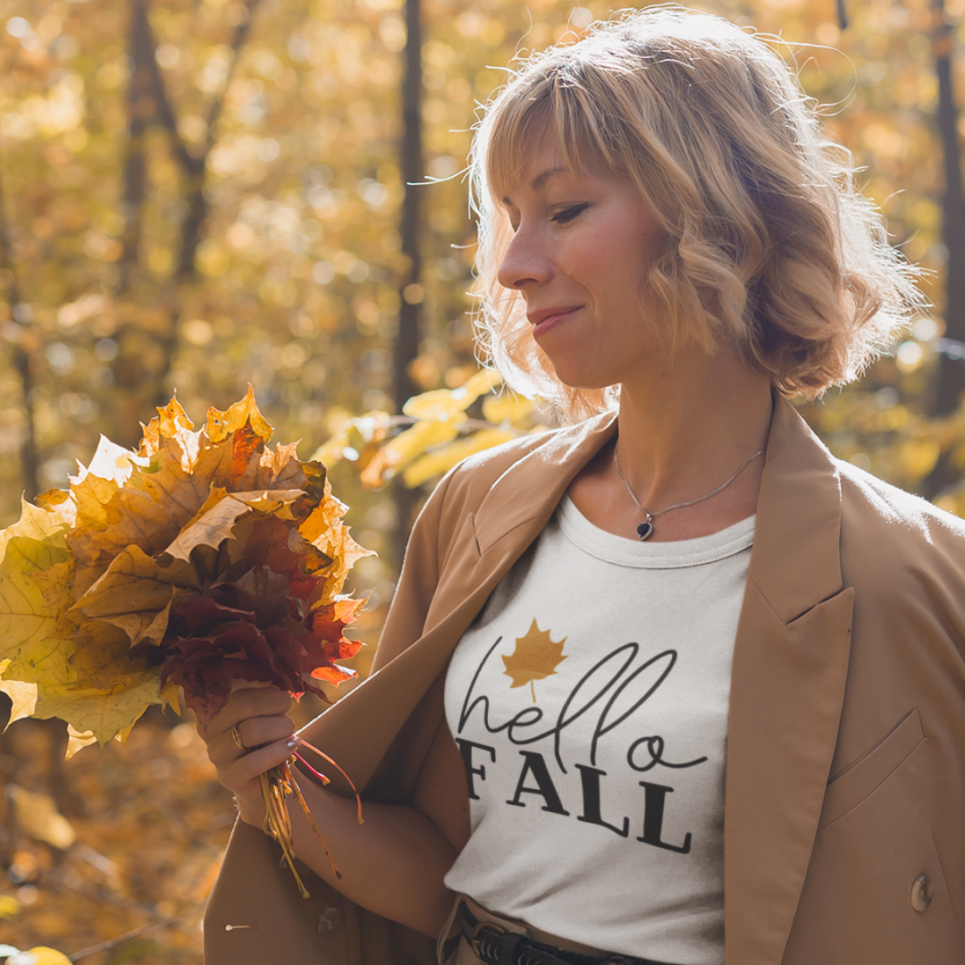White Hello Fall graphic T-shirt featuring an orange maple leaf and black lettering, displayed on autumn leaves.