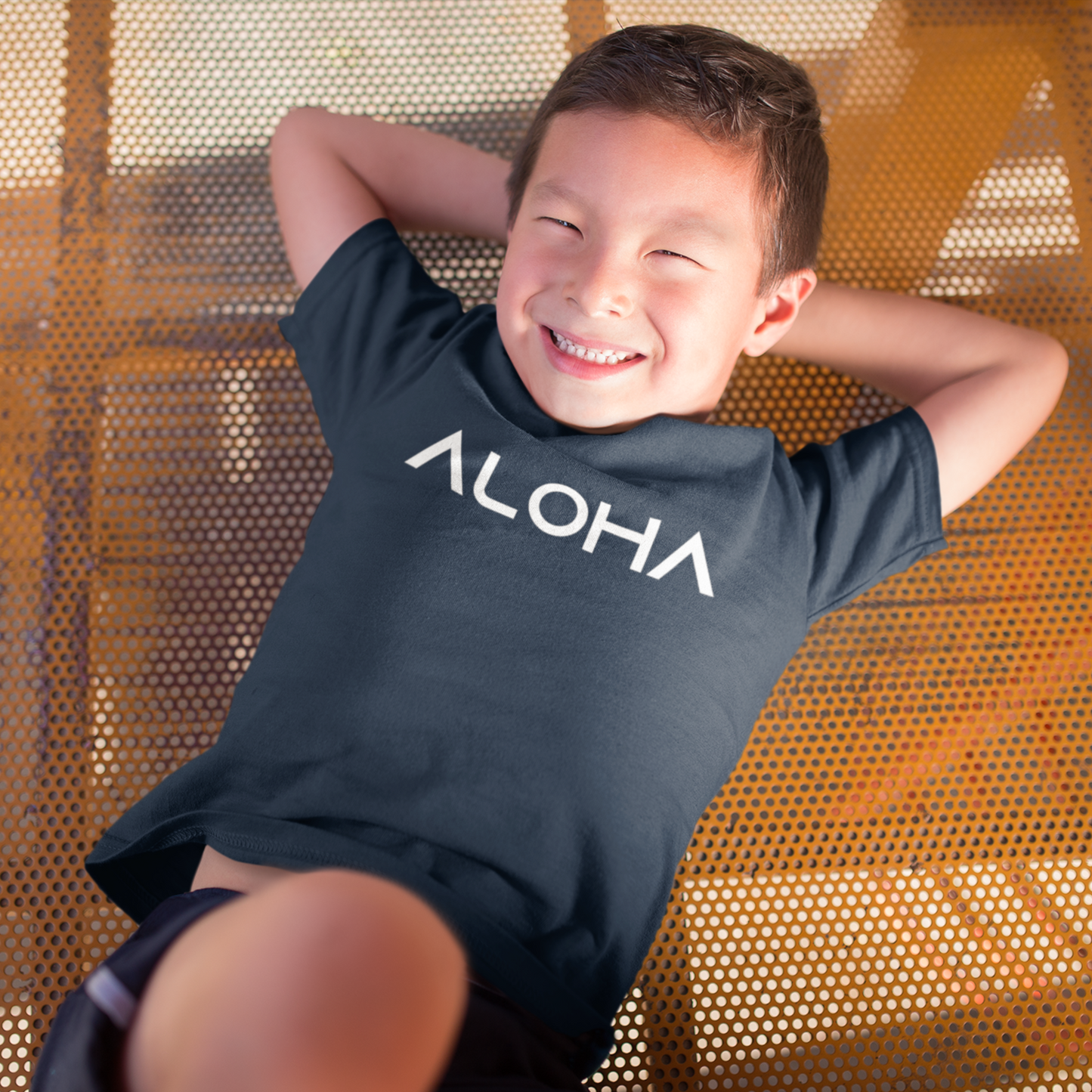 A young child wearing a black t-shirt that says "Aloha" in a white font, a Hawaiian-inspired shirt for kids. Available in different tropical colors and sizes
