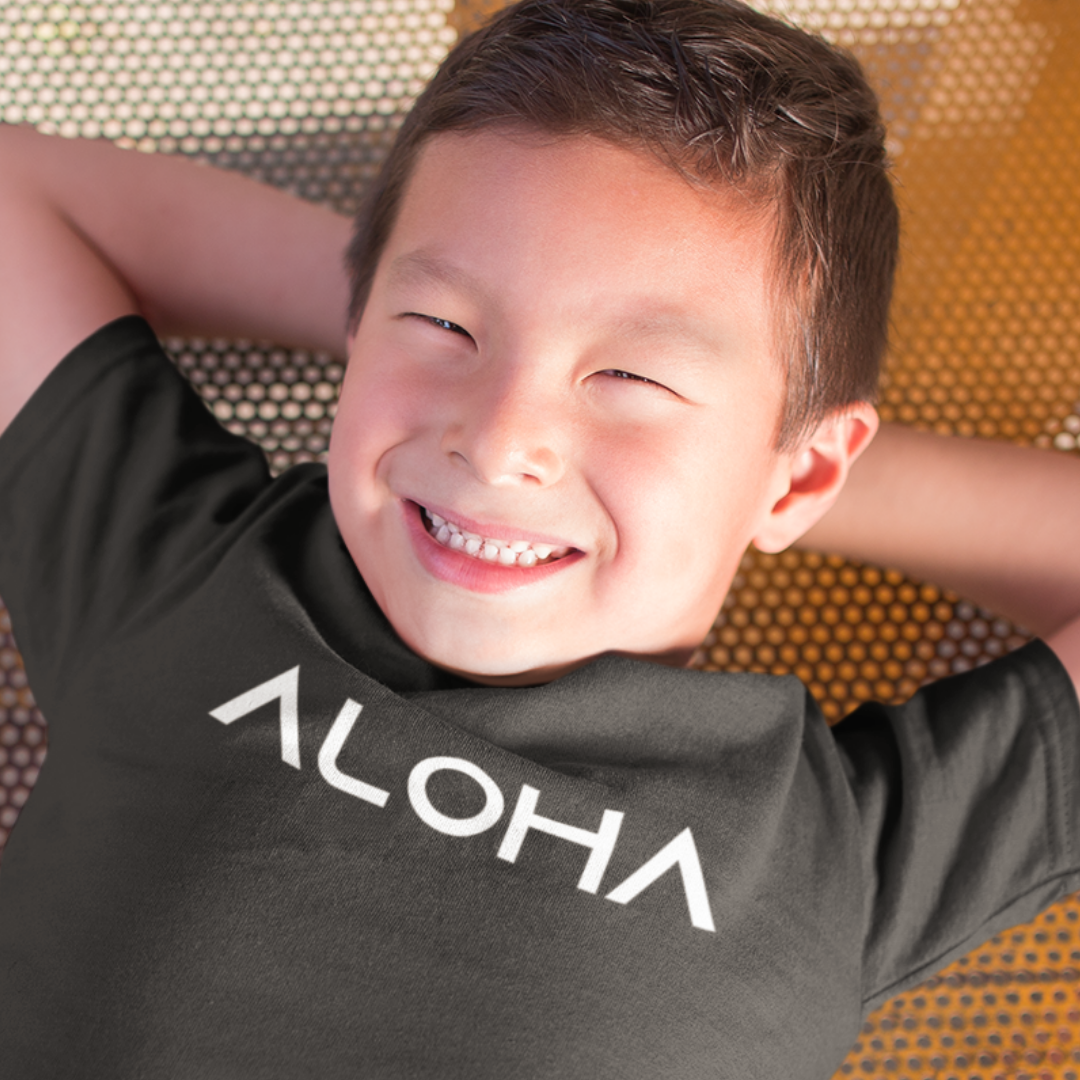 A young boy smiling while wearing a dark grey minimalist Kids Aloha Shirt from Simply Aloha On The Go.