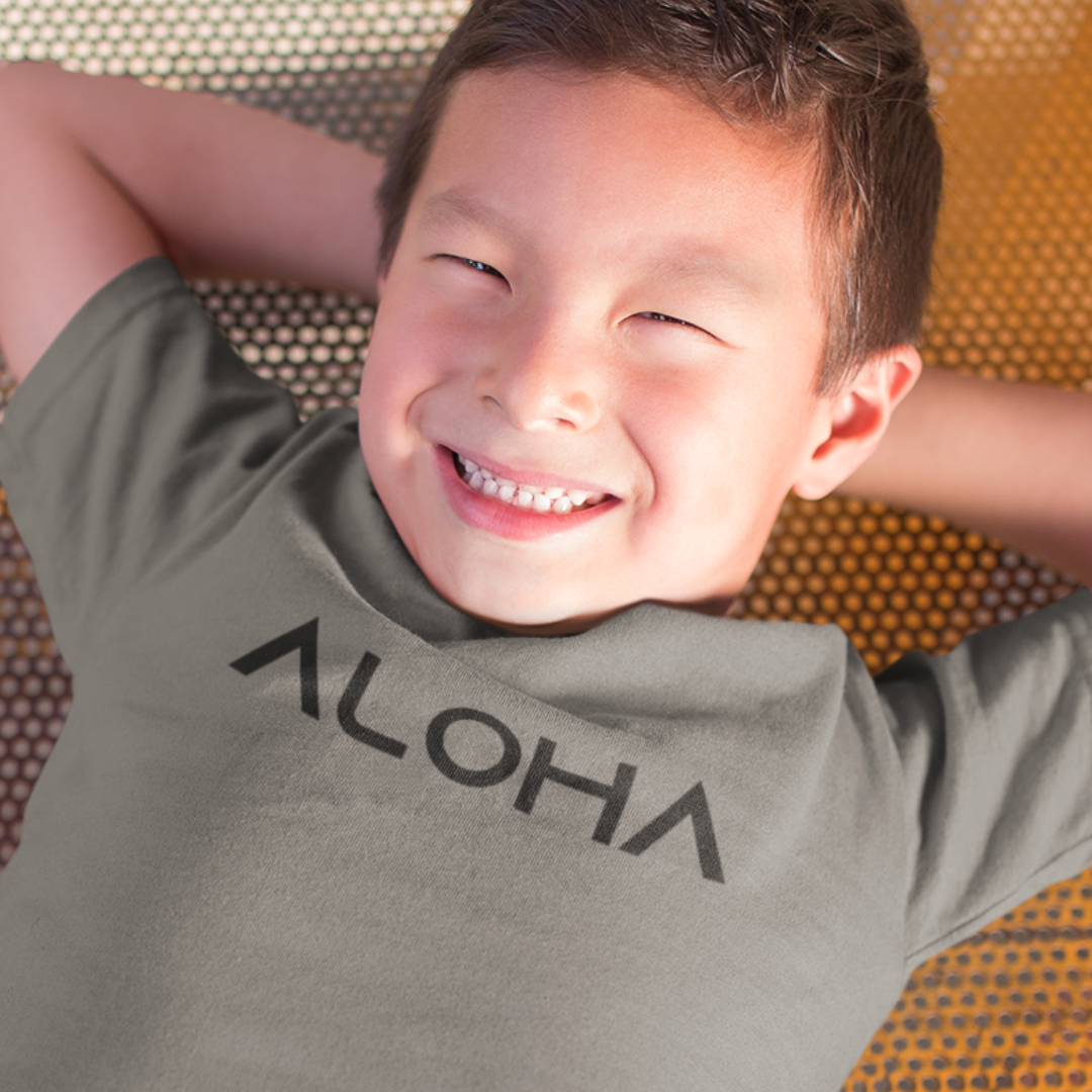 A young boy smiling while wearing a light grey minimalist Kids Aloha Shirt from Simply Aloha On The Go.